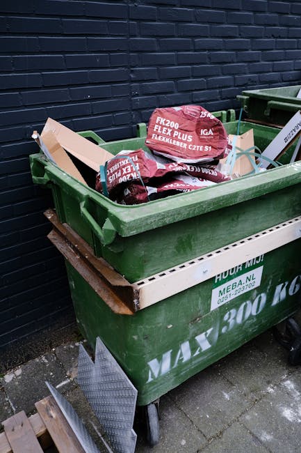 A green wheeled waste bin filled with several red and purple bags of outdoor garden or waste disposal material, stacked on top of a second larger green bin with a white label. The bins are positioned 